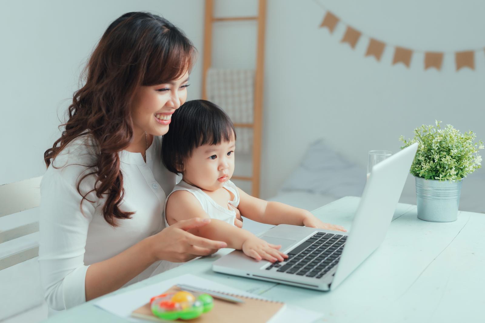 Working mother concept. Young woman working on laptop with her child from home